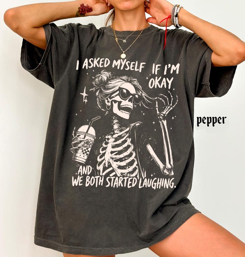 May include: Dark gray t-shirt featuring a skeleton graphic wearing sunglasses and holding a drink. The text on the shirt reads "I asked myself if I'm okay, and we both started laughing."