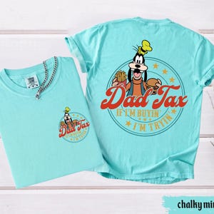 May include: Two chalky mint-colored t-shirts. One shirt has a small graphic on the front with the text "Dad Tax" and a cartoon character. The other shirt has a larger graphic on the back with the same text and character, plus the phrase "If I'm buyin', I'm tryin'."