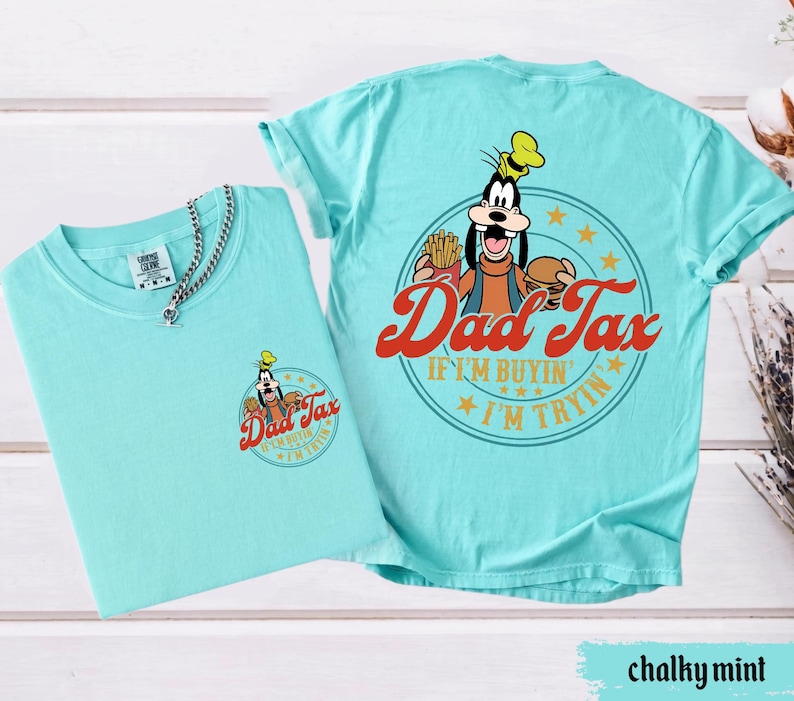 May include: Two chalky mint-colored t-shirts. One shirt has a small graphic on the front with the text "Dad Tax" and a cartoon character. The other shirt has a larger graphic on the back with the same text and character, plus the phrase "If I'm buyin', I'm tryin'."