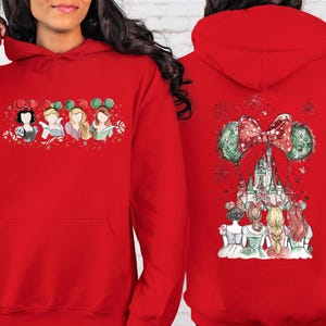 May include: Red hooded sweatshirt featuring a festive design. The front displays four cartoon princesses with mouse ears. The back showcases a castle with a bow and mouse ears. The design incorporates red, green, and white elements.