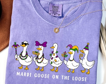 May include: A violet Comfort Colors t-shirt featuring a graphic of five cartoon geese in Mardi Gras attire. The geese are wearing sunglasses, bow ties, and party hats, with the text "MARDI GOOSE ON THE LOOSE" below. A gold necklace is draped across the shirt.