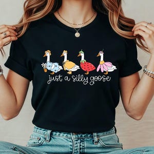 May include: Black t-shirt featuring a whimsical design of four geese dressed in various outfits, including a floral dress, a yellow cape, a strawberry costume, and a pink tutu. The text "just a silly goose" is written below the geese.