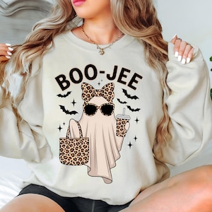 May include: Beige sweatshirt with a Halloween-themed graphic. The graphic features a ghost wearing sunglasses and a leopard print bow, holding a leopard print tote bag and a coffee cup. The text "BOO-JEE" is above the ghost.