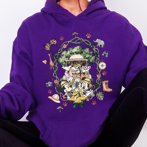 Peut inclure: Sweat &agrave; capuche violet avec un motif color&eacute; de personnages de dessins anim&eacute;s en tenue de safari. Le motif comprend une girafe, un &eacute;l&eacute;phant, une boussole et d'autres &eacute;l&eacute;ments sur le th&egrave;me du safari. Les personnages sont dispos&eacute;s autour d'un arbre central.
