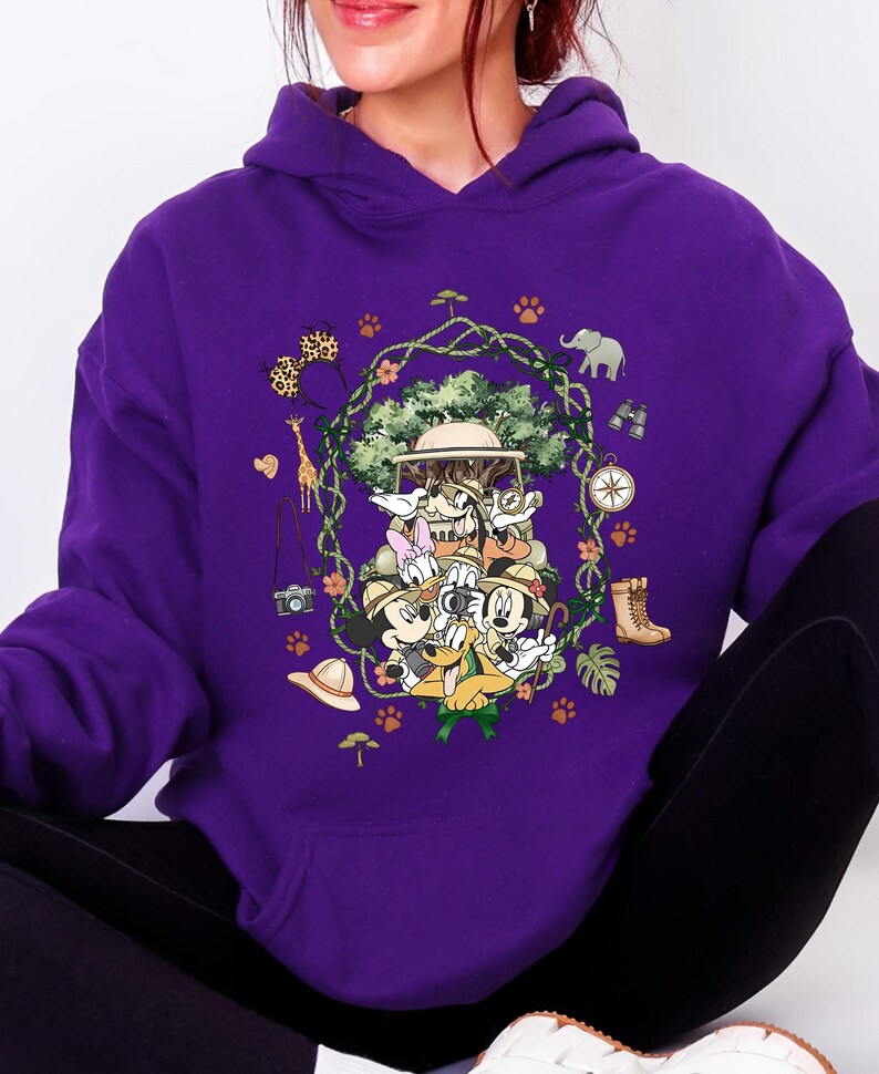 Peut inclure: Sweat &agrave; capuche violet avec un motif color&eacute; de personnages de dessins anim&eacute;s en tenue de safari. Le motif comprend une girafe, un &eacute;l&eacute;phant, une boussole et d'autres &eacute;l&eacute;ments sur le th&egrave;me du safari. Les personnages sont dispos&eacute;s autour d'un arbre central.