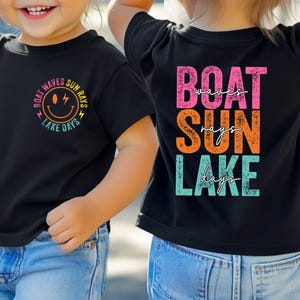 May include: Two black t-shirts with the text "BOAT WAVES SUN RAYS LAKE DAYS" and a smiley face graphic. The back of one shirt says "BOAT SUN LAKE" with "waves", "rays", and "days" underneath. The shirts are worn by a child.