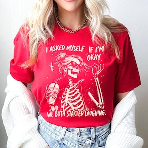 May include: Red t-shirt with a white graphic of a skeleton wearing sunglasses and holding a drink. The text on the shirt reads, "I ASKED MYSELF IF I'M OKAY, AND WE BOTH STARTED LAUGHING."
