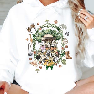 Peut inclure: Sweat &agrave; capuche blanc avec un motif safari color&eacute;. Le design pr&eacute;sente Mickey Mouse, Minnie Mouse, Donald Duck, Daisy Duck et Dingo en tenue de safari, entour&eacute;s d'illustrations li&eacute;es au safari, comme une girafe, des jumelles et une boussole.