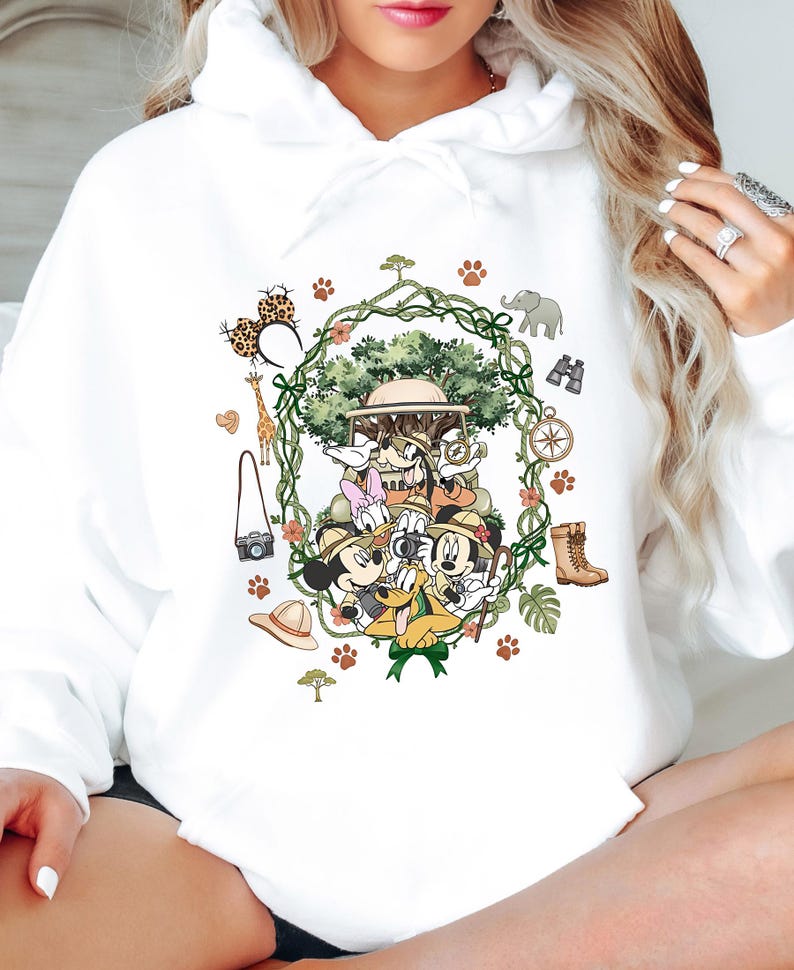 Peut inclure: Sweat &agrave; capuche blanc avec un motif safari color&eacute;. Le design pr&eacute;sente Mickey Mouse, Minnie Mouse, Donald Duck, Daisy Duck et Dingo en tenue de safari, entour&eacute;s d'illustrations li&eacute;es au safari, comme une girafe, des jumelles et une boussole.