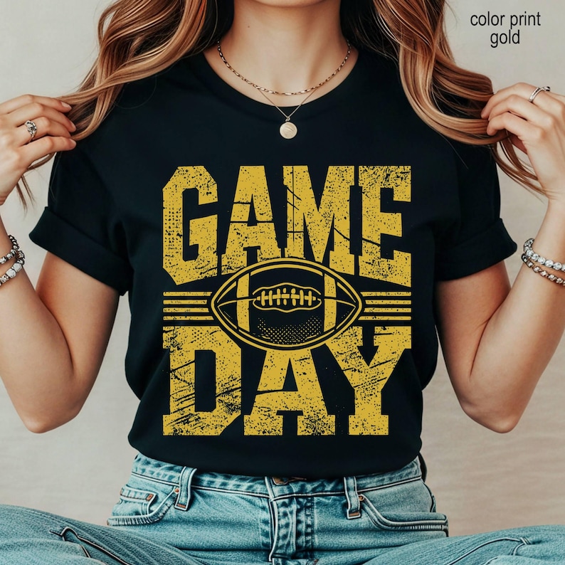 May include: A black t-shirt featuring the text GAME DAY in a distressed gold font. Centered between the words is a graphic of an American football. The design has a textured, worn appearance. The shirt is paired with blue denim jeans.