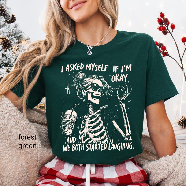 May include: Forest green t-shirt with a skeleton graphic wearing sunglasses and holding a beverage. The text on the shirt reads, "I asked myself if I'm okay, and we both started laughing."