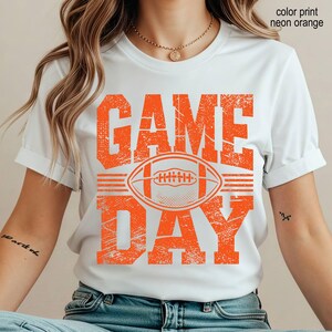 May include: A white short-sleeved t-shirt featuring a distressed neon orange graphic print that reads "GAME DAY" with a football illustration in the center. The text "color print neon orange" is visible in the upper right corner of the image.