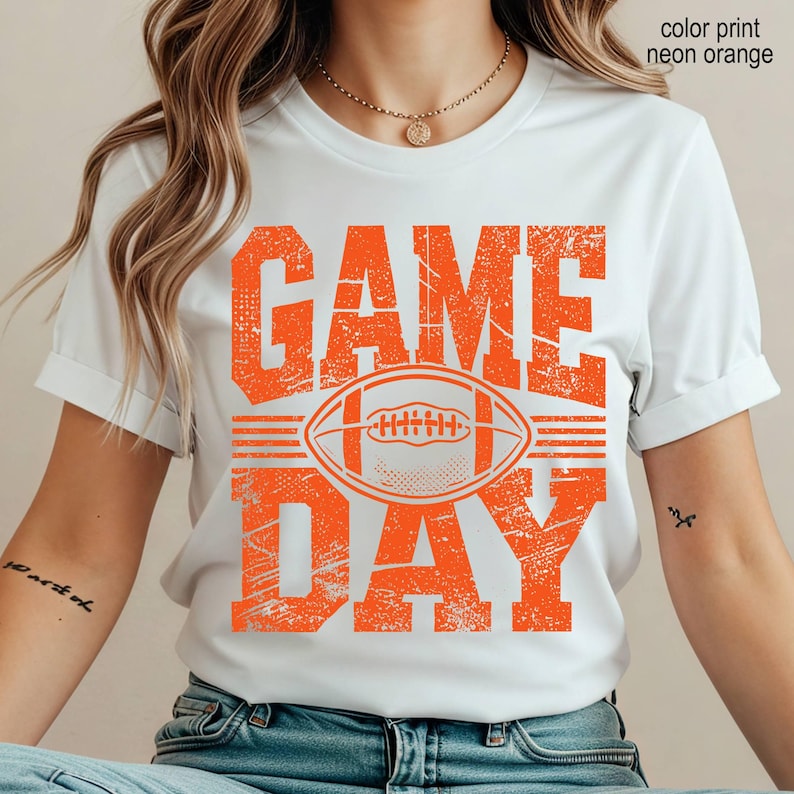 May include: A white short-sleeved t-shirt featuring a distressed neon orange graphic print that reads "GAME DAY" with a football illustration in the center. The text "color print neon orange" is visible in the upper right corner of the image.