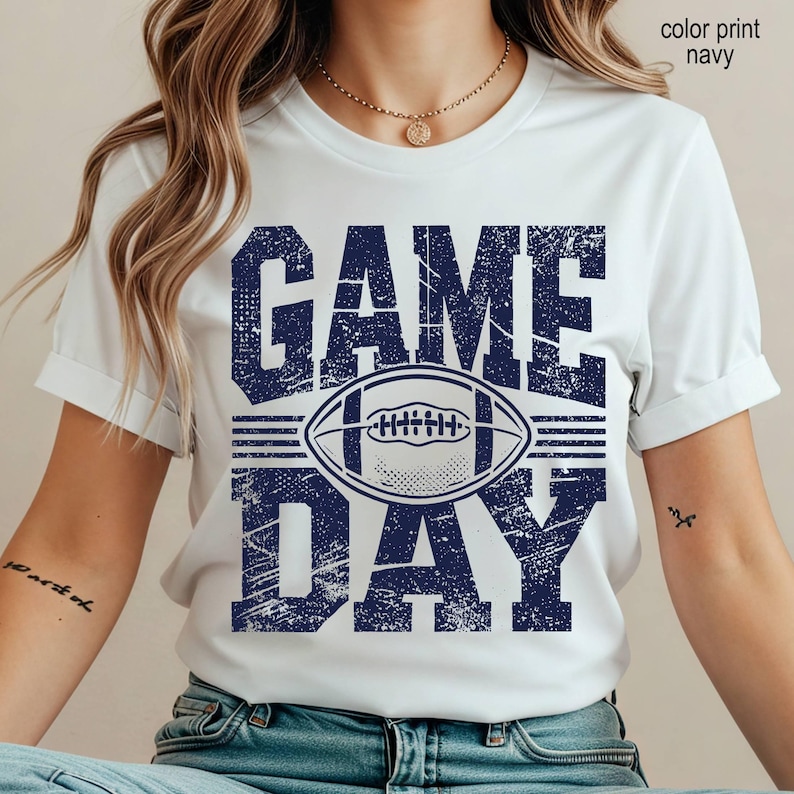May include: A white short-sleeved t-shirt featuring a distressed navy blue graphic print that reads "GAME DAY" with a football illustration centered between the words. The text and football design have a textured, weathered appearance.