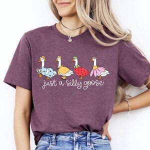 May include: A maroon t-shirt featuring a whimsical illustration of four geese, each adorned with unique outfits, including a floral dress, a yellow cape, a strawberry, and a pink skirt. The text "just a silly goose" is written below the geese.