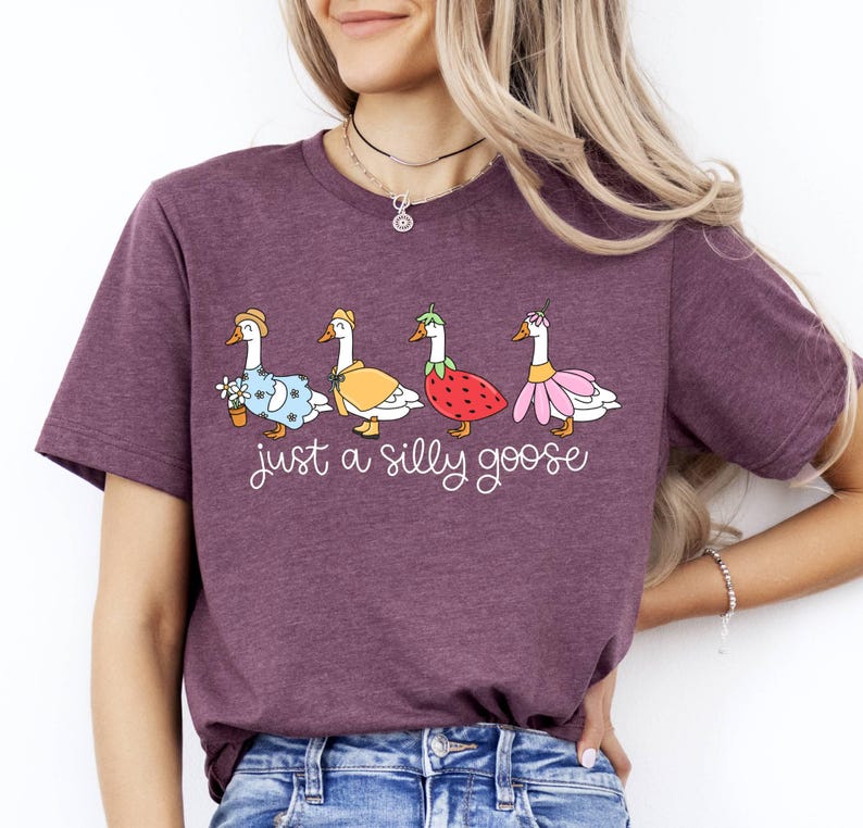 May include: A maroon t-shirt featuring a whimsical illustration of four geese, each adorned with unique outfits, including a floral dress, a yellow cape, a strawberry, and a pink skirt. The text "just a silly goose" is written below the geese.