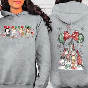May include: A light gray hoodie featuring a festive design. The front displays four cartoon princesses with Mickey Mouse ears and candy canes. The back showcases a castle scene with a large bow and Mickey Mouse ears.