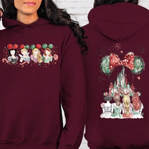 May include: Burgundy hoodie featuring a front graphic of four cartoon princesses with Mickey Mouse ears and candy canes. The back of the hoodie displays a castle with a Minnie Mouse bow and ears, and fireworks.