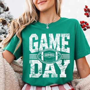 Puede incluir: Camiseta verde con las palabras "GAME DAY" en letras blancas desgastadas y un gr&aacute;fico de f&uacute;tbol americano blanco. La camiseta es de cuello redondo y manga corta.