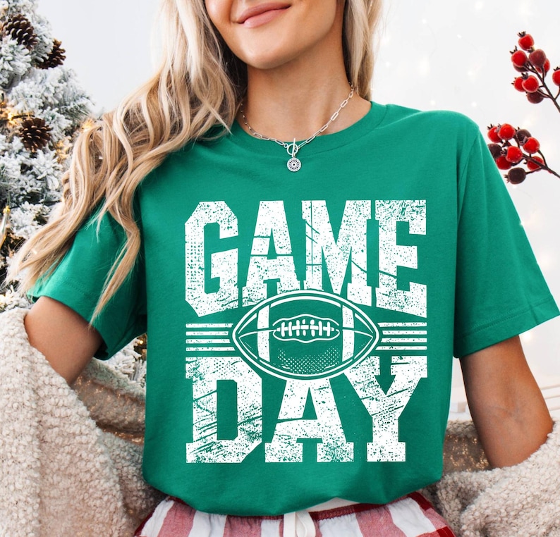 Puede incluir: Camiseta verde con las palabras "GAME DAY" en letras blancas desgastadas y un gr&aacute;fico de f&uacute;tbol americano blanco. La camiseta es de cuello redondo y manga corta.