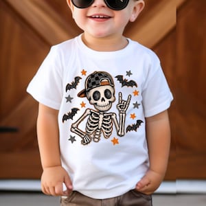 May include: A white short-sleeve t-shirt featuring a graphic of a skeleton wearing a black and white checkered baseball cap. The skeleton is making a hand gesture surrounded by small black bats and orange and gray stars printed on the front.