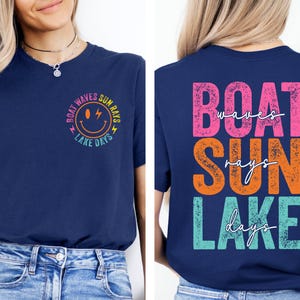 May include: A navy blue t-shirt with a vibrant graphic design. The front displays a smiley face and the words "Boat Waves Sun Rays Lake Days". The back of the shirt has the words "Boat Waves Sun Rays Lake Days" in a stacked layout.
