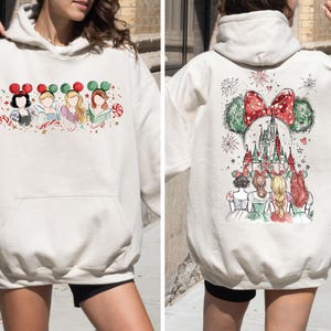 May include: Cream-colored hoodie featuring a watercolor design of Disney princesses on the front and back. The front displays four princesses with festive headbands. The back showcases a castle and fireworks.