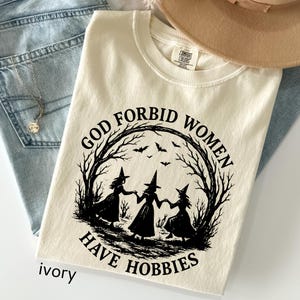May include: Ivory-colored t-shirt with the text "GOD FORBID WOMEN HAVE HOBBIES" in black. The graphic features three witch silhouettes holding hands inside a tree branch circle with bats. The shirt is folded on top of blue jeans.