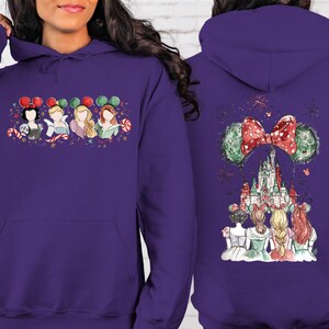 May include: A purple hooded sweatshirt featuring a front graphic of four cartoon princesses wearing holiday-themed mouse ears. The back of the sweatshirt displays a castle scene with a large bow and mouse ears.