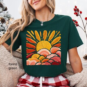May include: A forest green t-shirt featuring an embroidered design of a sun rising over layered, colorful clouds. The sun is yellow, with orange and red rays. The clouds are shades of pink, orange, and red. The text "forest green" is on the left side.
