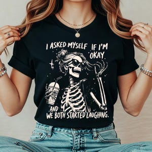 May include: Black t-shirt featuring a skeleton wearing sunglasses and holding a drink. The text on the shirt reads, "I asked myself if I'm okay, and we both started laughing."