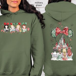 May include: Military green hoodie featuring a front graphic of Disney princesses with candy canes and mouse ears. The back displays a castle, fireworks, and a large bow with mouse ears. A festive design.