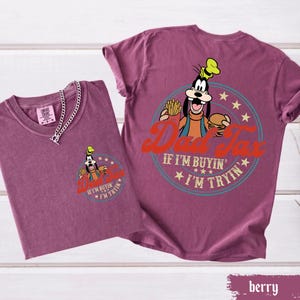 May include: Two berry-colored t-shirts. One shirt has a small graphic of Goofy holding fries and a burger, with text that reads "IF I'M BUYIN' I'M TRYIN'". The other shirt has a larger graphic of Goofy with the text "Dad Tax" and "IF I'M BUYIN' I'M TRYIN'".