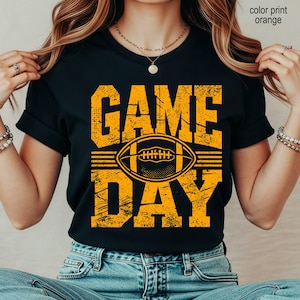 May include: A black short-sleeved t-shirt featuring a distressed orange graphic print that reads GAME DAY with a football illustration in the center. The text and football design have a textured, worn appearance on the front of the shirt.