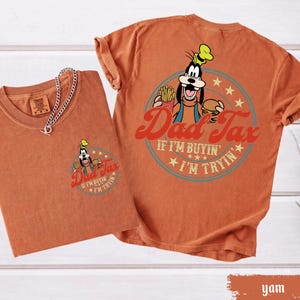 May include: Two orange-brown t-shirts. One shirt has a small graphic of Goofy holding fries and a burger with the text "Dad Tax". The other shirt has a larger version of the same graphic and text. The text reads "If I'm buyin', I'm tryin'."