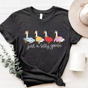 May include: Dark gray t-shirt featuring a whimsical illustration of four geese dressed in various outfits, including a floral dress, a yellow cape, a strawberry costume, and a pink flower dress. The text "just a silly goose" is written below the geese in a cursive font.