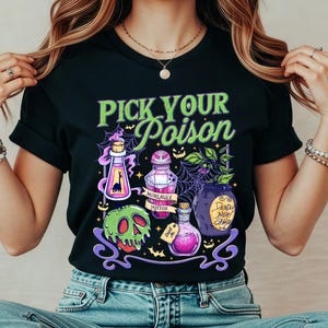 May include: Black t-shirt with a Halloween-themed graphic. The design features the text "PICK YOUR POISON" in green, along with illustrations of potion bottles, a skull, and a spiderweb. The shirt is made of a soft material.