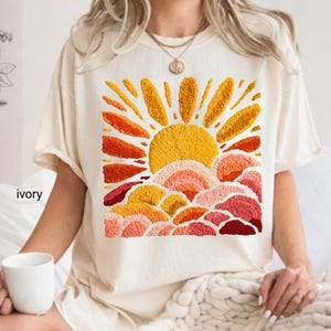 May include: Ivory-colored t-shirt featuring an embroidered design of a sun rising over layered clouds. The sun is a bright yellow, with orange and red rays. The clouds are in shades of orange, pink, and red.