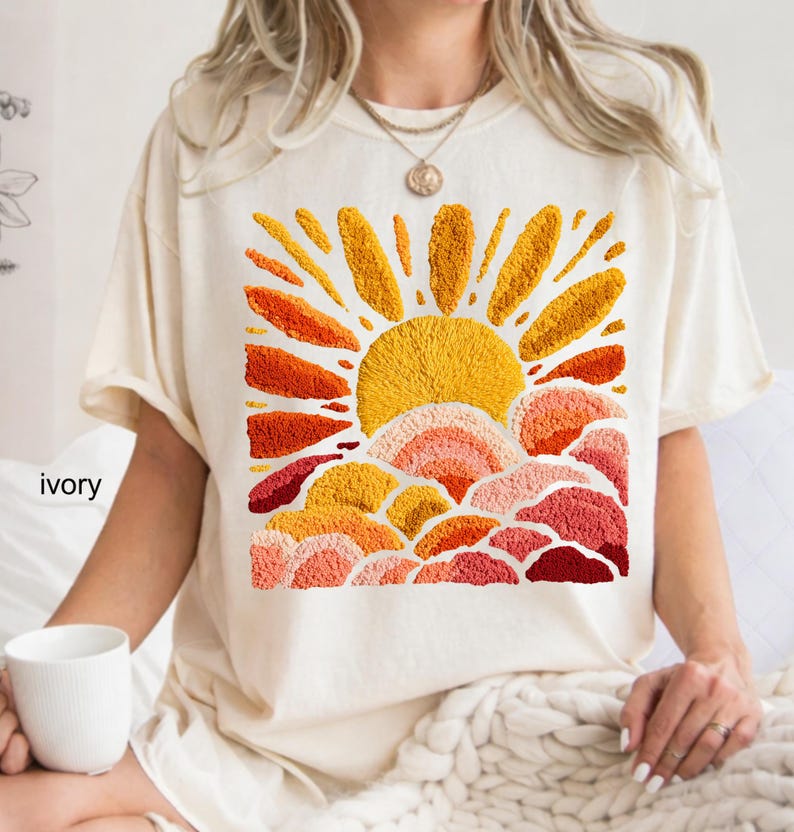 May include: Ivory-colored t-shirt featuring an embroidered design of a sun rising over layered clouds. The sun is a bright yellow, with orange and red rays. The clouds are in shades of orange, pink, and red.