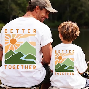 May include: White t-shirts with a mountain and sun graphic, and the words "BETTER TOGETHER" in orange. The shirts are worn by a person and a child, suggesting a matching set.