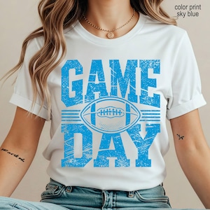 May include: A white short-sleeve t-shirt featuring a distressed sky blue graphic print. The design includes the text "GAME DAY" in large block letters with a football illustration centered between the words. The text "color print sky blue" is visible.