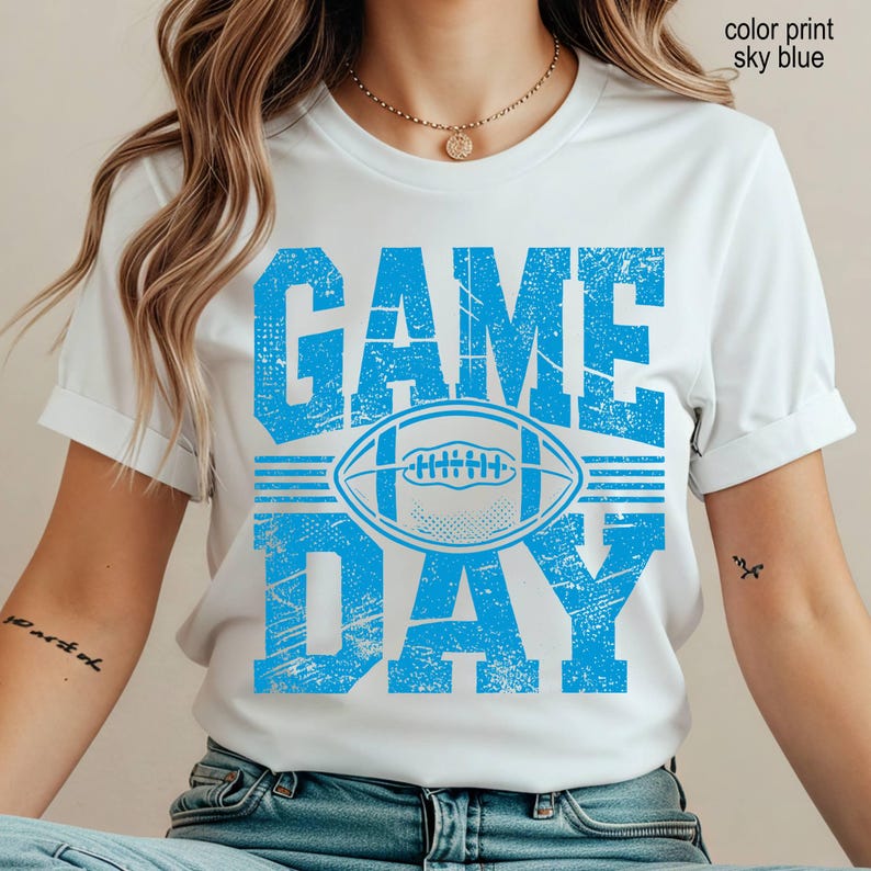 May include: A white short-sleeve t-shirt featuring a distressed sky blue graphic print. The design includes the text "GAME DAY" in large block letters with a football illustration centered between the words. The text "color print sky blue" is visible.