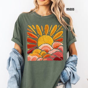 May include: Moss green t-shirt featuring an embroidered design of a sun with orange and yellow rays, and layered clouds in shades of orange, pink, and red. The word "moss" is printed in black on the upper right. The shirt has rolled-up sleeves.