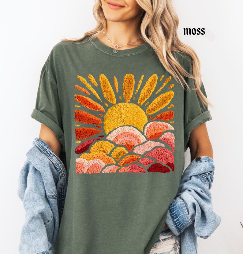 May include: Moss green t-shirt featuring an embroidered design of a sun with orange and yellow rays, and layered clouds in shades of orange, pink, and red. The word "moss" is printed in black on the upper right. The shirt has rolled-up sleeves.