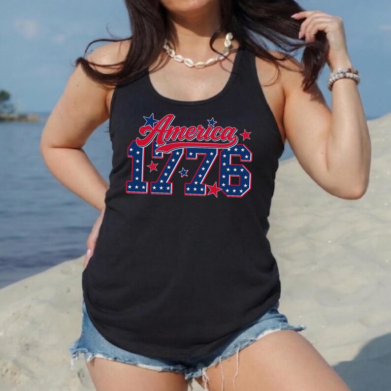Op de afbeelding: Zwarte tanktop met het woord "America" in rode letters en het nummer "1776" in blauw met witte sterren. De tanktop wordt gecombineerd met een spijkerbroek. De foto is buiten bij het water gemaakt.