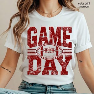May include: A white short-sleeved t-shirt featuring a distressed maroon graphic print that reads GAME DAY with a football illustration in the center. The text color print is maroon. The shirt has a crew neckline and a standard fit.