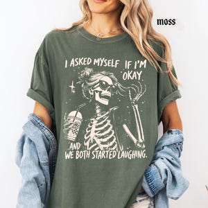 May include: Moss green t-shirt with a skeleton graphic wearing sunglasses and holding a drink. The text on the shirt reads, "I asked myself if I'm okay, and we both started laughing."