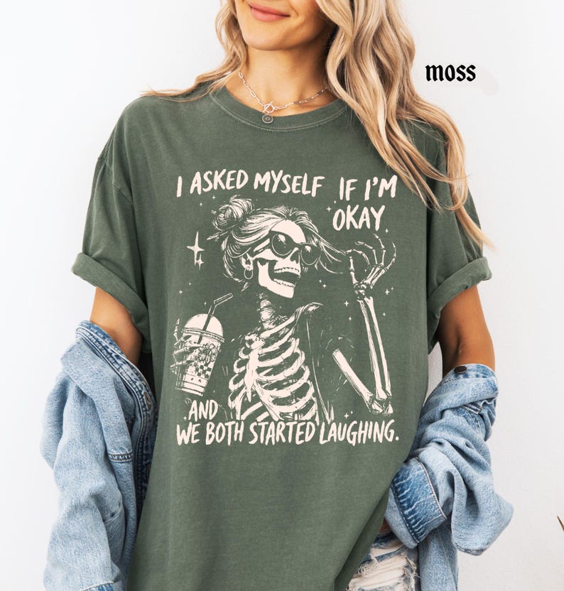 May include: Moss green t-shirt with a skeleton graphic wearing sunglasses and holding a drink. The text on the shirt reads, "I asked myself if I'm okay, and we both started laughing."