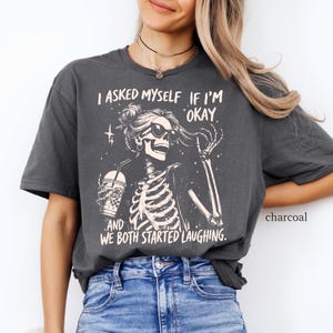 May include: Charcoal-colored t-shirt featuring a skeleton graphic wearing sunglasses and holding a drink. The text reads, "I asked myself if I'm okay, and we both started laughing."