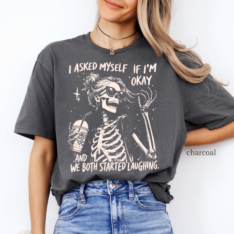 May include: Charcoal-colored t-shirt featuring a skeleton graphic wearing sunglasses and holding a drink. The text reads, "I asked myself if I'm okay, and we both started laughing."